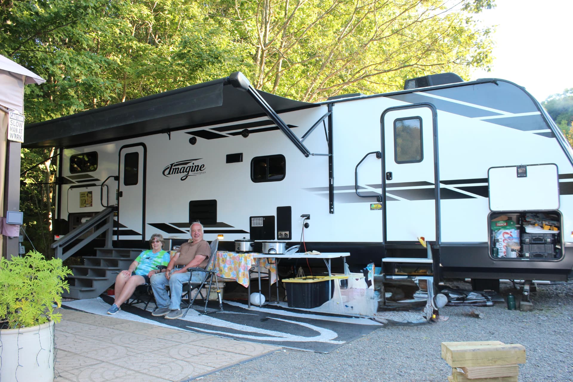 A couple relaxing in camp chairs under the awning of their travel trailer, with a table of slow cookers beside them