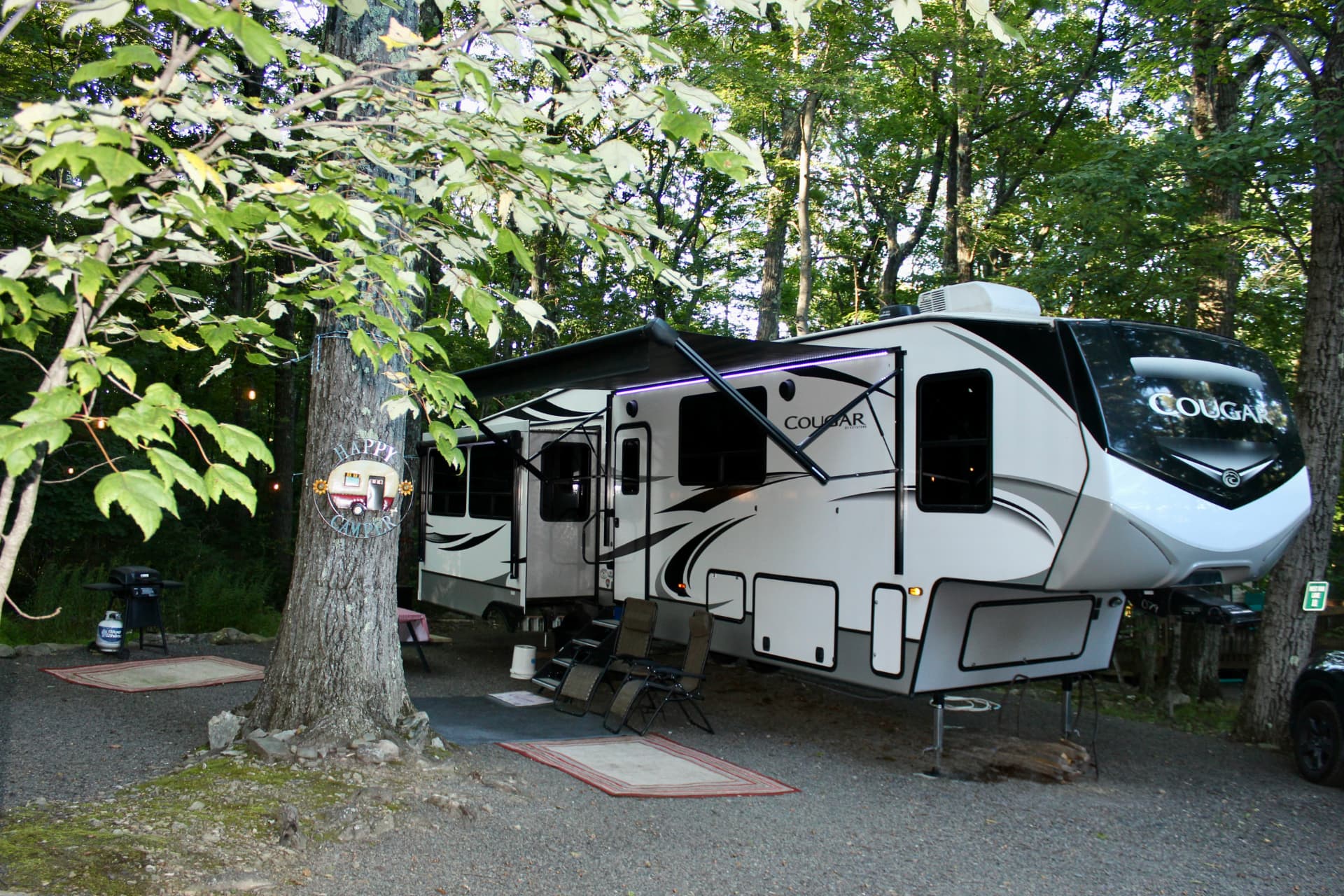 A fifth-wheel RV with its awning open at a shaded gravel campsite, two camping chairs and a grill set up beside it